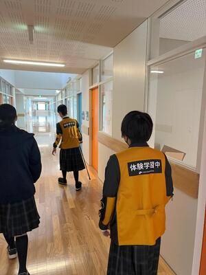 学校の廊下にて、片麻痺体験や高齢者体験をして歩く生徒たち