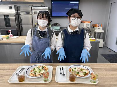 普通科２年１組の生徒がトレイの上の料理を紹介しています。トレイの上にはチーズが溶けた熱々のピザとフルーツがいっぱい入ったジュレが乗っています。